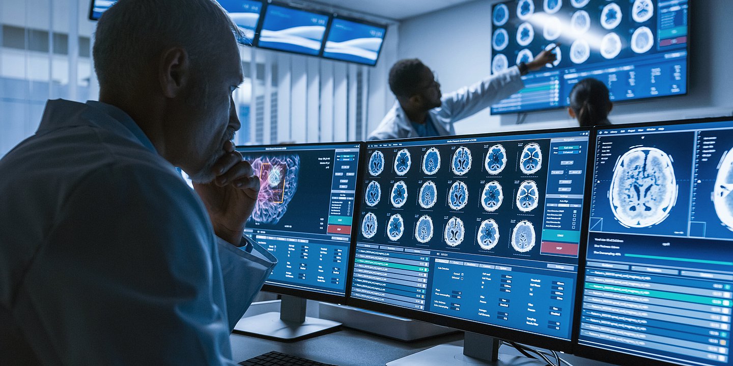 Ein Arzt sitzt am Schreibtisch vor 3 Monitoren. Auf ihnen zu sehen sind verschiedene Hirnscans.