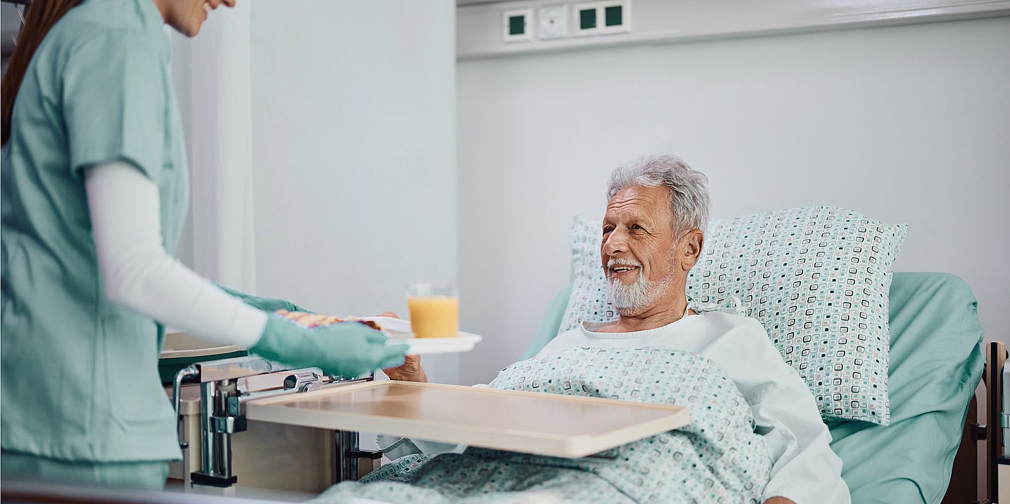 Ein hochbetagter Mann bekommt Frühstück mit einem Orangensaft an sein Krankenhausbett getragen.
