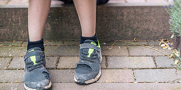 Abgenutzte, vorne aufgerissene Turnschuche an den Füßen eines Kindes, stehend auf Pflastersteinen