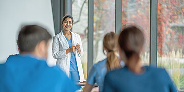 Das Bild zeigt eine Frau in wei&szlig;en Kittel und darunter blauer Krankenhaus-Kleidung, die eine Gruppe unterichtet. Die Gruppe tr&auml;gt ebenfalls blaue Klinik-Kleidung und wendet dem Beobachter den R&uuml;cken zu. Im Hintergrund ist eine gro&szlig;e Fensterfront.