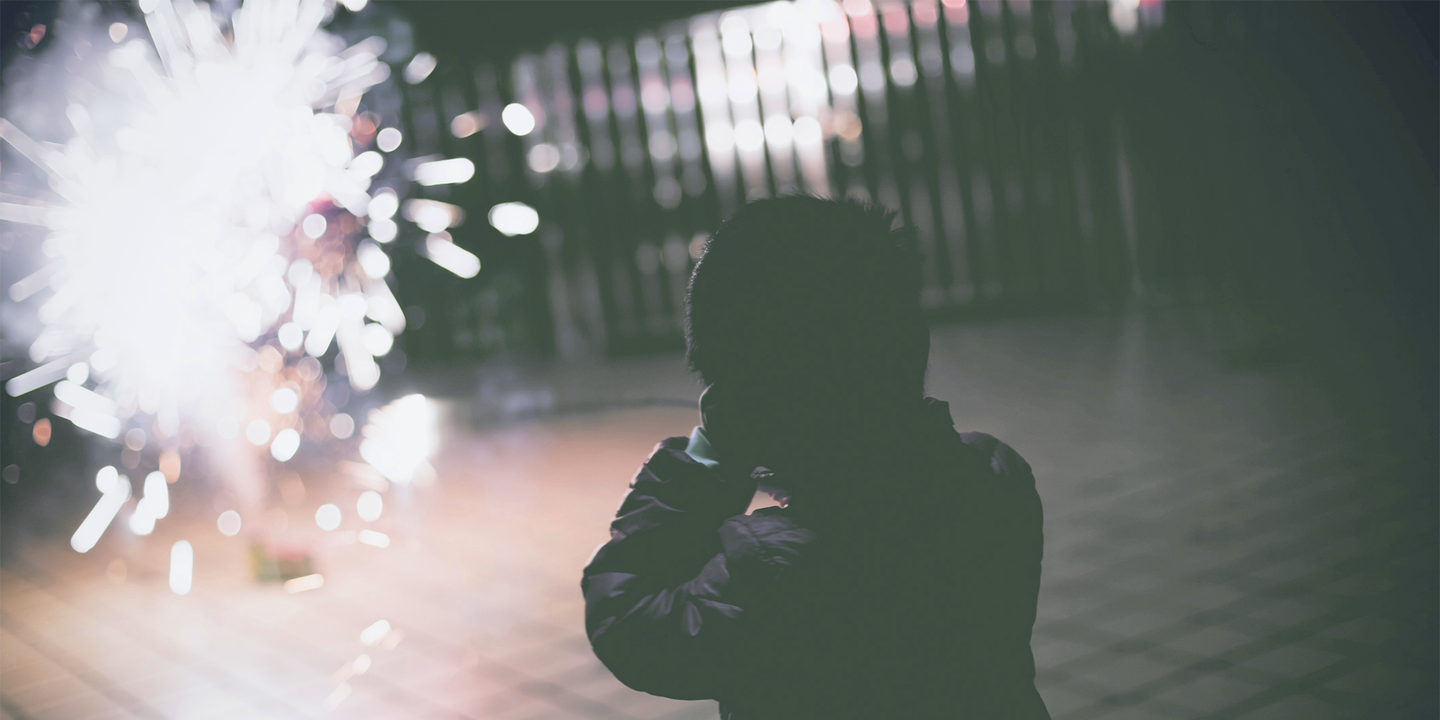 Foto: Ein Mensch hält sich die Ohren zu, während etwas entfernt eine Silvesterrakete explodiert.