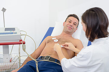 Ein m&auml;nnlicher Patient liegt mit freiem Oberk&ouml;rper auf einer Behandlungsliege. Eine Mitarbeiterin legt die Elektroden f&uuml;r das EKG an.