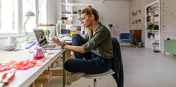 Eine Frau sitzt im Schneidersitz auf einem B&uuml;rostuhl vor einem Laptop. Um sie herum liegen kreative Materialien, wie Krepppapier und ein Stanzer.