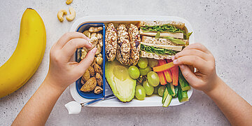 Auf einem Tisch liegt eine Brotbox mit belegtem Vollkornbrot, Gem&uuml;se, N&uuml;ssen und M&uuml;slikeksen. Zwei Kinderh&auml;nde greifen nach M&ouml;hrensticks. Links daneben liegen eine Banane und ein paar Cashew-N&uuml;sse.