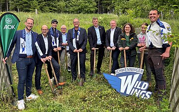 Eine Gruppe von Menschen steht vor einem Wald um B&auml;ume zu pflanzen.