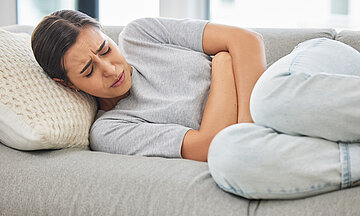 Eine junge Frau liegt seitlich mit angezogenen Beinen auf einem Sofa. Die Arme sind &uuml;ber den Bauch verschr&auml;nkt, das Gesicht ist schmerzlich verzogen, die Augen geschlossen.