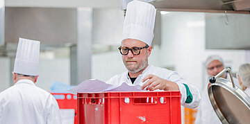 Koch mit wei&szlig;er M&uuml;tze arbeitet in einer Gro&szlig;k&uuml;che, weitere Mitarbeitende im Hintergrund.