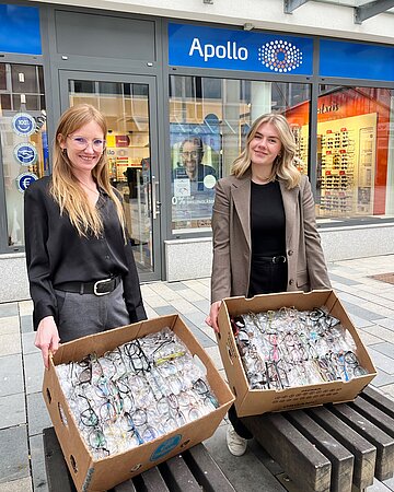 Fanny Liebich und Livia Ha&szlig;ler stehen vor dem Schaufenster von Apollo-Optik in B&ouml;blingen und zeigen zwei Kisten voller eingesammelter Brillen.