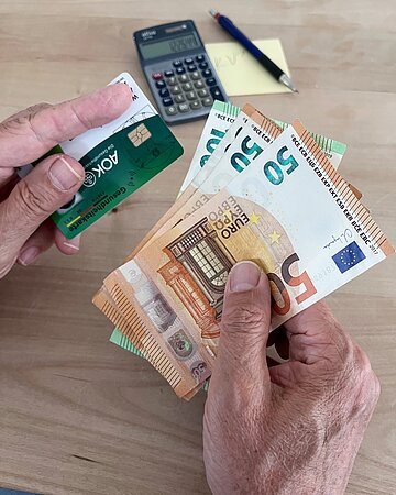 Ein paar H&auml;nde vor einem Taschenrechner und einem Zettelblock: Die linke Hand h&auml;lt eine AOK-Versichertenkarte, die rechte einige Euro-Scheine.