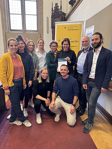 Im Foto stehend von links nach rechts in der oberen Reihe: Christina Schub mit Greta Wirth und Bernadette H&uuml;lsmann (alle vom Projekt "M&uuml;nchen &ndash; gesund vor Ort"), Dr. Verena Lindacher (Abteilungsleitung Stadtteilgesundheit), Beatrix Zurek (Gesundheitsreferentin), Verena Dietl (B&uuml;rgermeisterin), Martin Felber (Direktor der Direktion M&uuml;nchen, AOK Bayern) und Andreas Nagel (Referent Lebenswelten, AOK Bayern) sowie in der unteren Reihe Lena Donhauser und Sven von Braum&uuml;ller (beide: Projekt "M&uuml;nchen &ndash; gesund vor Ort")