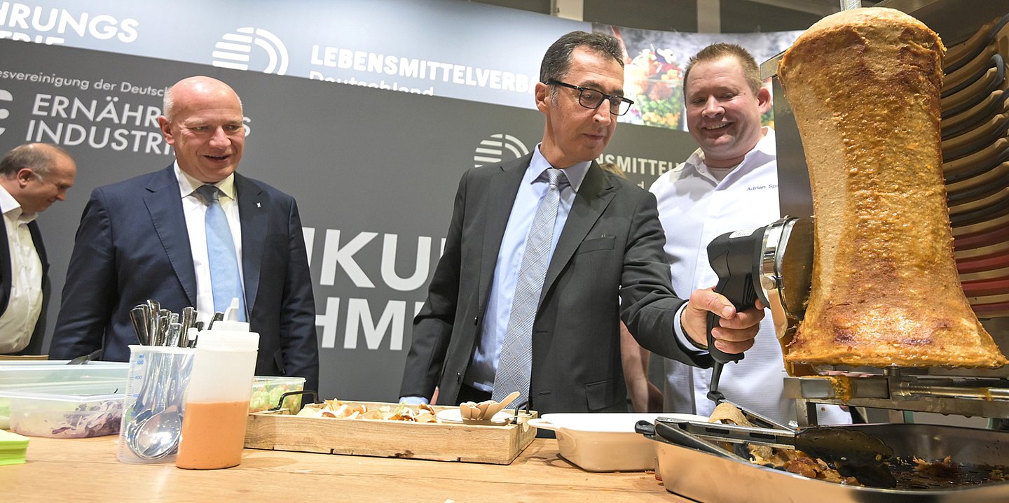 Foto: Bundeslandwirtschaftsminister Cem Özdemir und Berlins Regierender Bürgermeister Kai Wegner stehen an einem Messestand. Özdemir schneidet Fleisch von einem veganen Dönerspieß. 