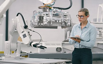 Eine Frau mit Tablet steht in einem Labor und begutachtet einen Roboterarm.