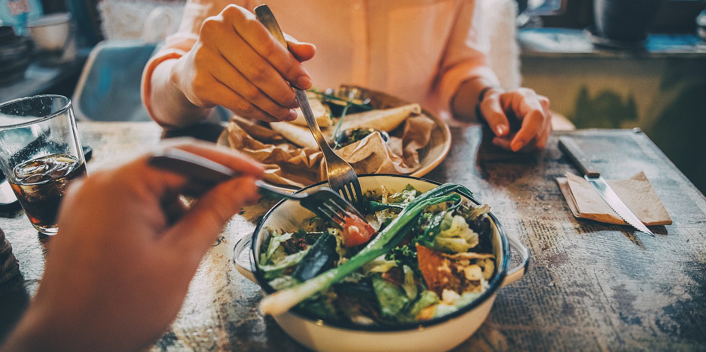 Foto: Zwei Teller mit Essen stehen auf einem Tisch – auf dem einen ist mehr Gemüse, auf dem anderen mehr teighaltiges Essen. Die Hände zweier Menschen mit Gabeln essen von dem Teller mit Gemüse. 