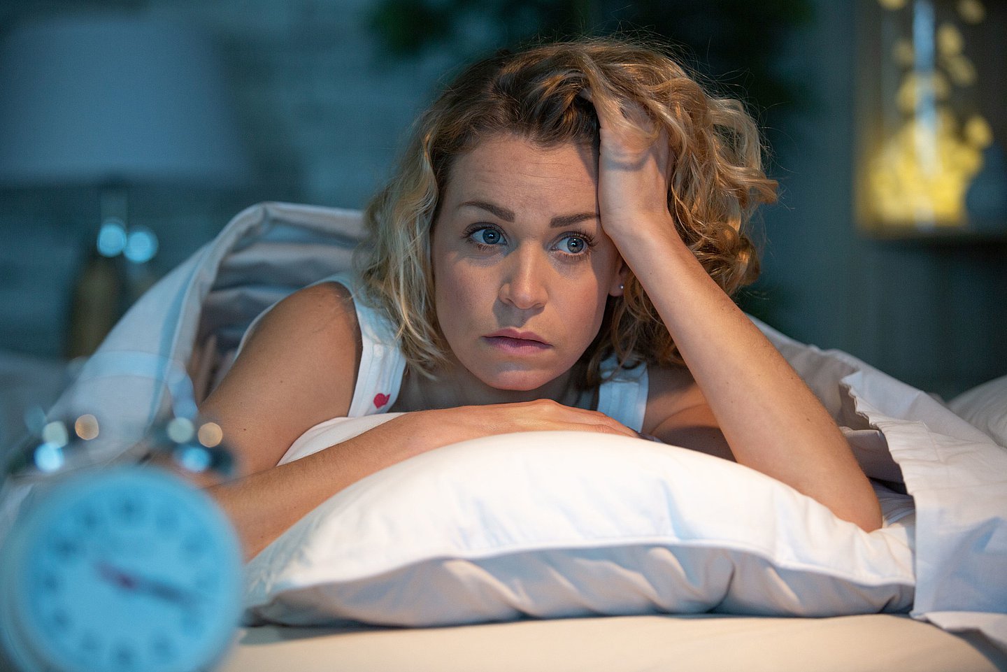 Foto zeigt eine Frau nachts in ihrem Bett liegend, die sich mit der linken Hand den Kopf auf ihrem Kopfkissen abstützt und etwas genervt schaut, weil sie nicht schlafen kann.