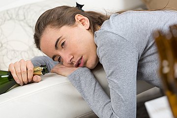 Komasaufen in Schleswig-Holstein Eine Jugendliche sitzt auf dem Boden, lehnt sich auf/gegen ein Sofa, die Augen halb geschlossen, ins Leere blickend, den Kopf aufgestützt auf der linken Hand, die auf der Sitzfläche des Sofas liegt. Die rechte hält eine geöffnete Flasche.