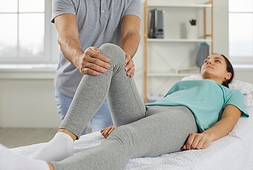 Heilmitteltherapien in Schleswig-Holstein Eine junge Frau liegt auf einer Behandlungsliege beim Physiotherapeuten. Das linke Bein liegt ausgestreckt, das rechte Bein ist aufgestellt und wird auf Kniehöhe vom Behandler umfasst.