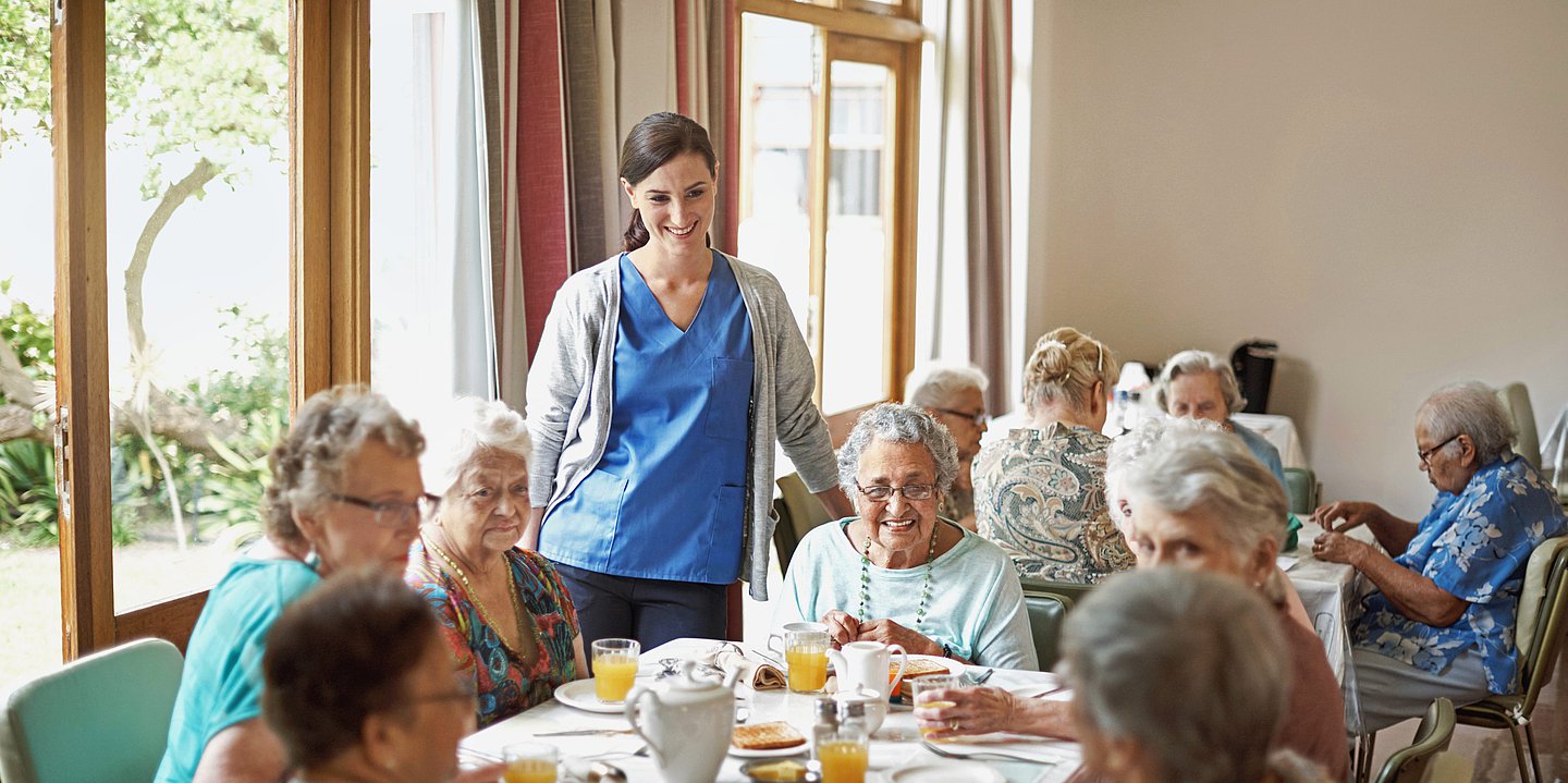 Das Foto zeigt eine zufrieden, freundlich lächelnde Pflegerin mit blaugrüner Dienstkleidung und einer hellgrauen Strickjacke mitten im Speisesaal an der Kaffeetafel. Im Hintergrund sind vier weitere Seniorinnen an einem Tisch zu sehen. im Speisesaal eines Seniriorenheimes stehend am Tisch mit sechs Senirionnen 