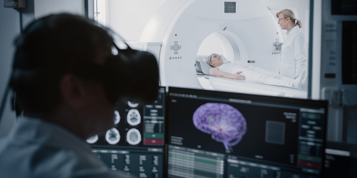 Blick eines Forschenden vor einem Bildschirm mit Bildern eines Gehirns. Im Hintergrund ist ein MRT zu sehen.