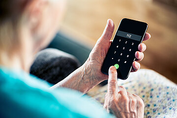 Eine sitzende &auml;ltere Frau im Schlafanzug h&auml;lt ein Handy in der Hand und dr&uuml;ckt gerade den Notruf 112.