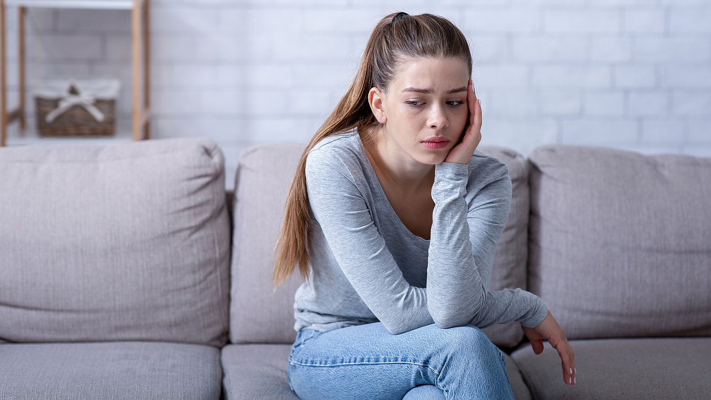 Eine junge Frau sitzt auf einem Sofa, sie hat den Kopf auf die linke Hand gestützt und schaut traurig.