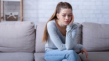Eine junge Frau sitzt auf einem Sofa, sie hat den Kopf auf die linke Hand gest&uuml;tzt und schaut traurig.