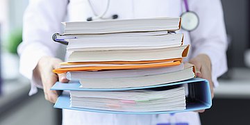 Bürokratie in der Gesundheitswirtschaft Foto eines Mediziners, der einen Stapel Akten vor sich trägt