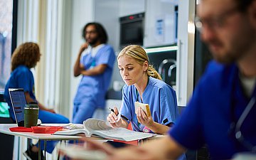 Eine Krankenpflegerin sitzt an einem Schreibtisch und arbeitet in einem Buch. Sie hat ein Sandwich in der Hand. Vor und hinter ihr sind weitere Krankenpflegende, entweder am Tisch oder stehend in eine Unterhaltung vertieft.