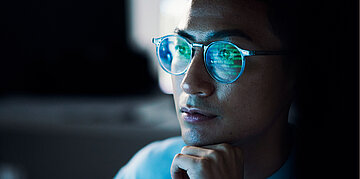 Bild eines Mannes mit Brille, der in einen Bildschirm blickt. In seiner Brille spiegelt sich der schwarze Bildschirm mit gr&uuml;ner Schrift