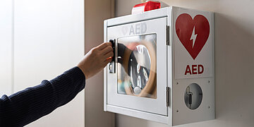 Eine wei&szlig;e Box mit einem gro&szlig;en roten Herz und einem wei&szlig;en Blitz darin h&auml;ngt an der Wand. Unter dem Herz steht "AED". Die Box hat eine T&uuml;r, eine Person macht diese gerade auf.