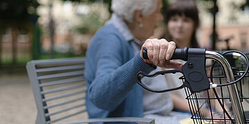 Im Vordergrund steht ein Rollator, den eine Frau am Lenker h&auml;lt. Sie sitzt unscharf im Hintergrund auf einer Bank und neben ihr sitzt ein junges M&auml;dchen.