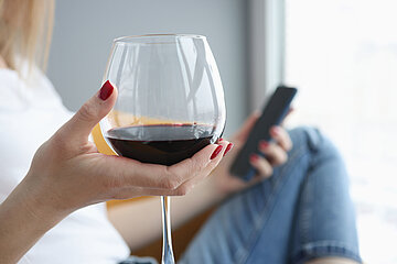Eine Frau in Nahaufnahme sitzt mit dem Handy und einem Glas Rotwein auf der Couch.