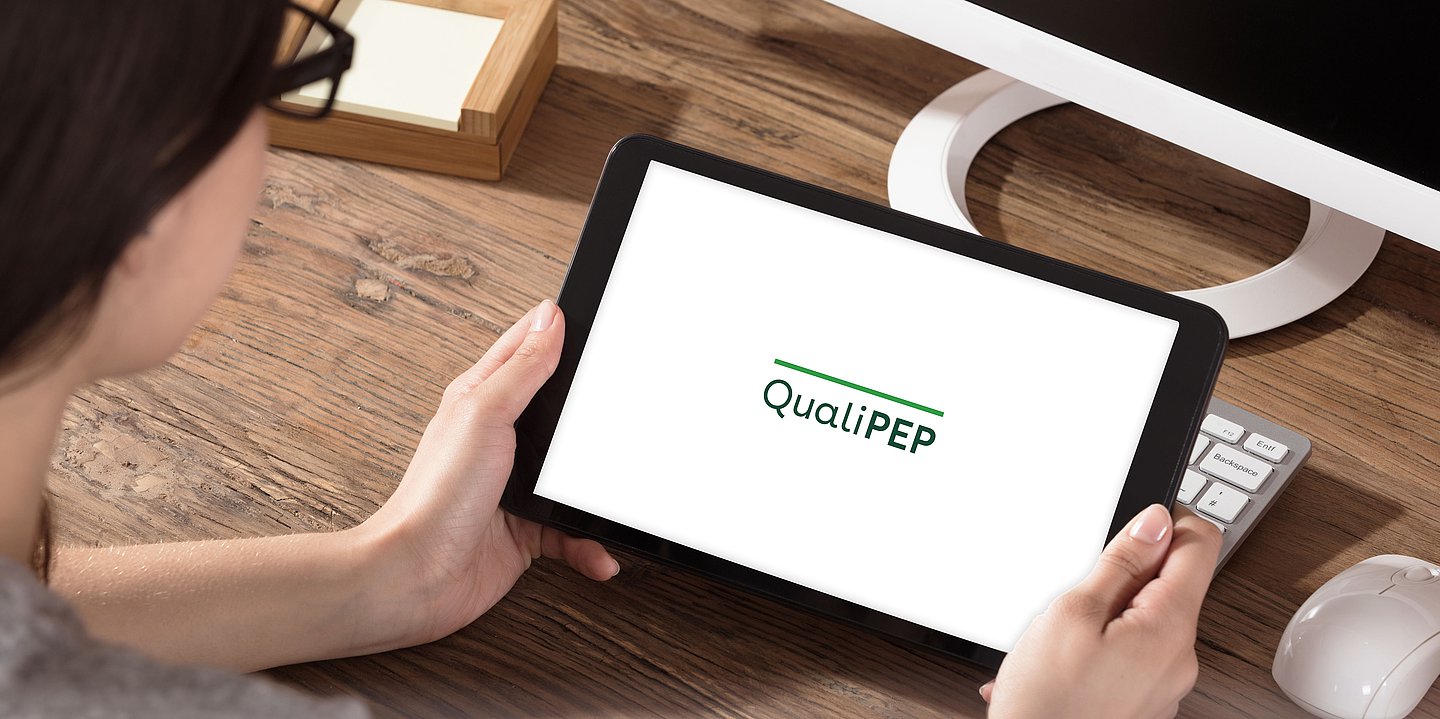 Foto: Eine Frau sitzt am Schreibtisch, hält ein Tablet in der Hand. Auf dem Tablet ist das QualiPEP-Logo zu sehen.