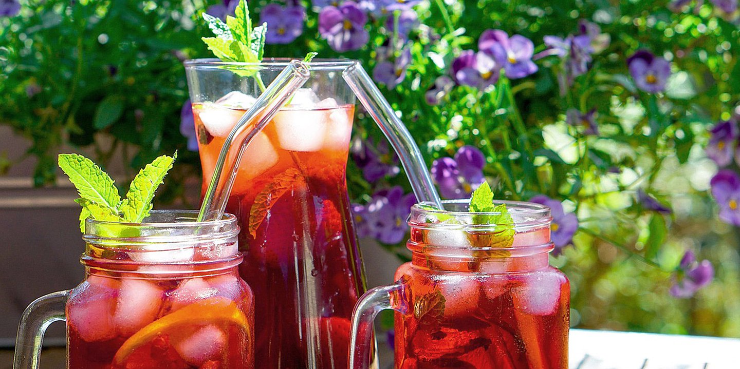 Auf einem Tisch im Freien stehen drei Gläser mit rotem Saft und Eiswürfeln, sowie Strohhalmen und Minzdekoration.