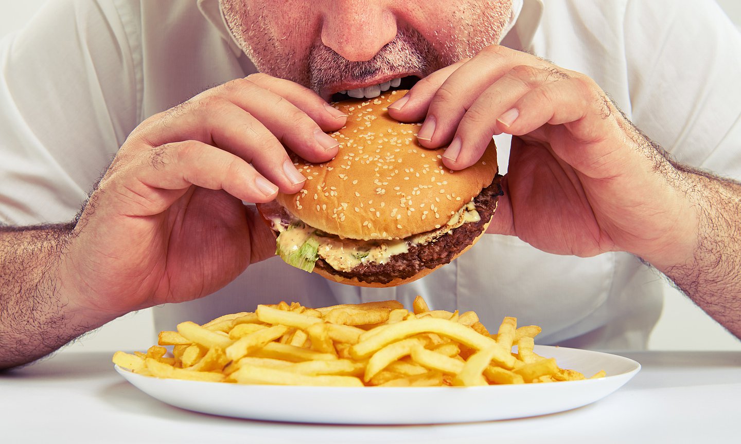 Eine männliche Person sitzt am Tisch, vor sich einen großen Teller mit Pommes frites, und beißt gerade in einen Burger, den er mit beiden Händen hält.