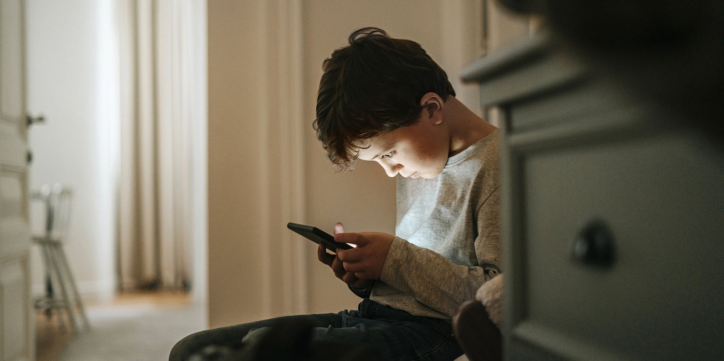 Ein Junge sitzt in einem Flur hinter einer Kommode und starrt mit gesenktem Kopf auf ein Smartphone.