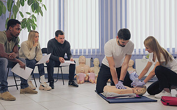 In einem Erste-Hilfe-Kurs schauen drei Erwachsene, die auf St&uuml;hlen sitzen, zu, wie eine junge Frau und ein Mann an einer Puppe Beatmung und Herzmassage &uuml;ben.