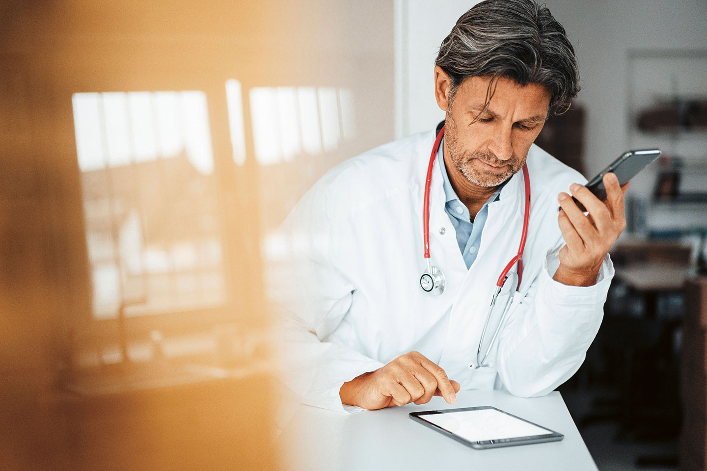 Foto: Ein Mann im Arztkittel tippt auf einem Tablet, hält ein Smartphone in der Hand und trägt ein Stethoskop um den Hals.