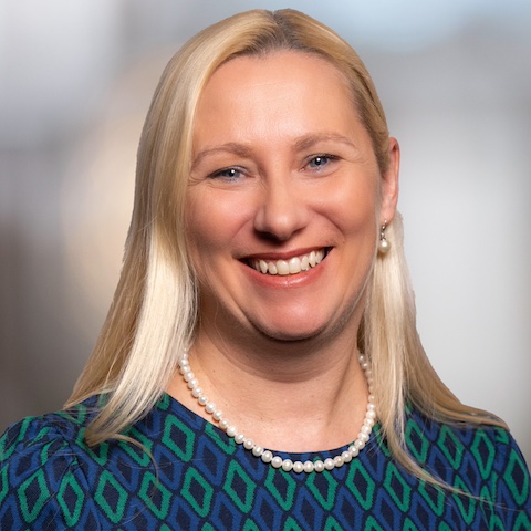 Diana Stolz Porträtfoto zeigt Diana Stolz mit blonden, schulterlangem Haar und weißer Perlenkette. Um den Hals deutet sich fdas dunkelblau-grün-gemusterte Kleid.