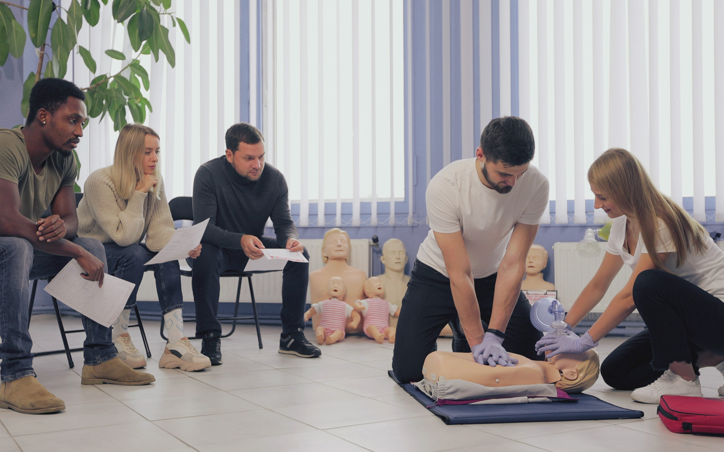 In einem Erste-Hilfe-Kurs schauen drei Erwachsene, die auf Stühlen sitzen, zu, wie eine junge Frau und ein Mann an einer Puppe Beatmung und Herzmassage üben.