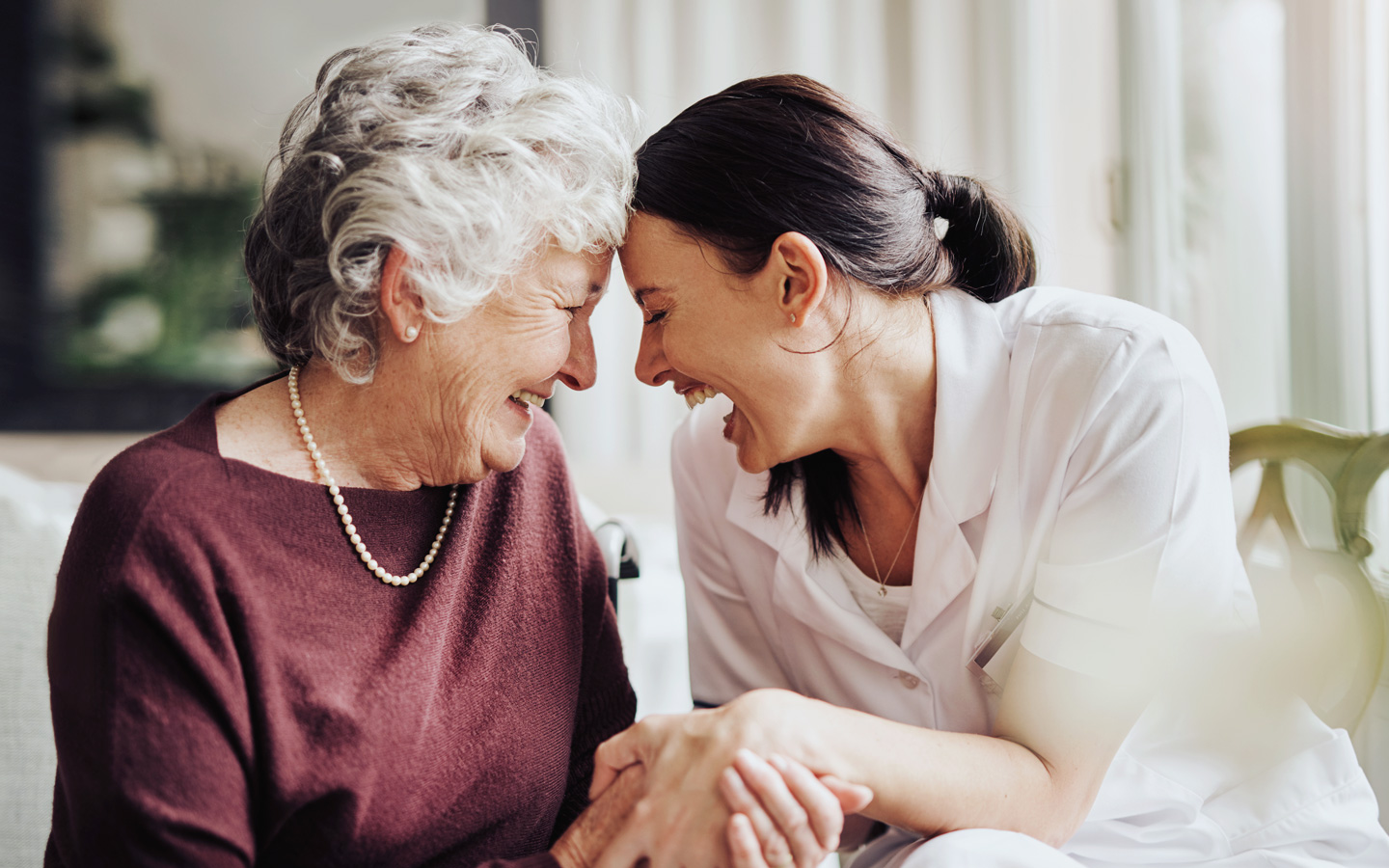 Eine ältere zu pflegende Frau sitzt gemeinsam mit einer Pflegekraft und lacht.