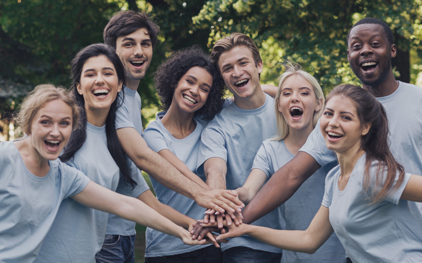 Mehrere Pflegekräfte verschiedener Herkunft und Hautfarben stehen als Gruppe und lachen gemeinsam und halten ihre Hände zusammen.