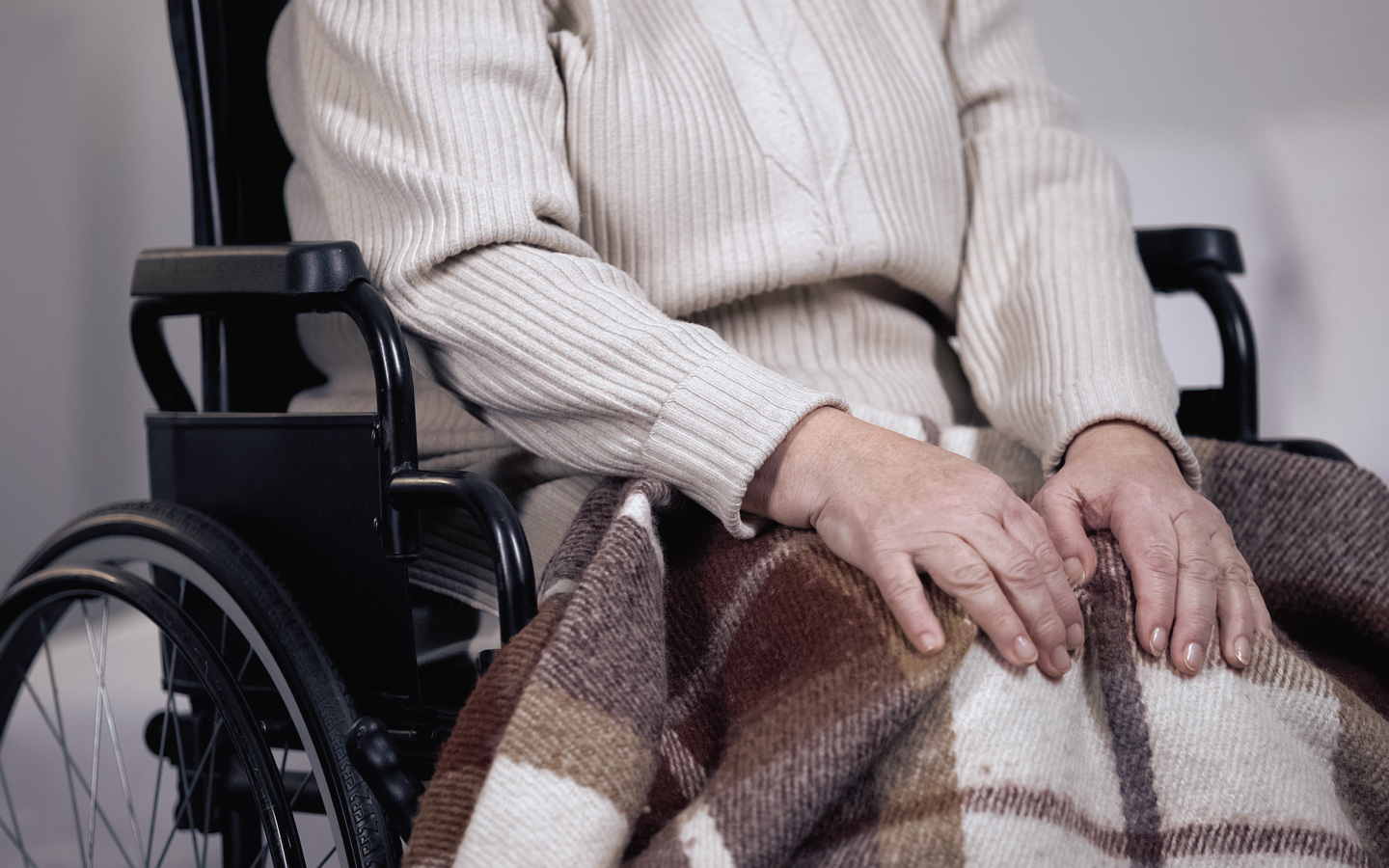 Auf dem Bild sieht man eine angezogene Person im Rollstuhl mit einem hellen Pullover und einer Wolldecke über den Knien. Offenbar wartet sie.