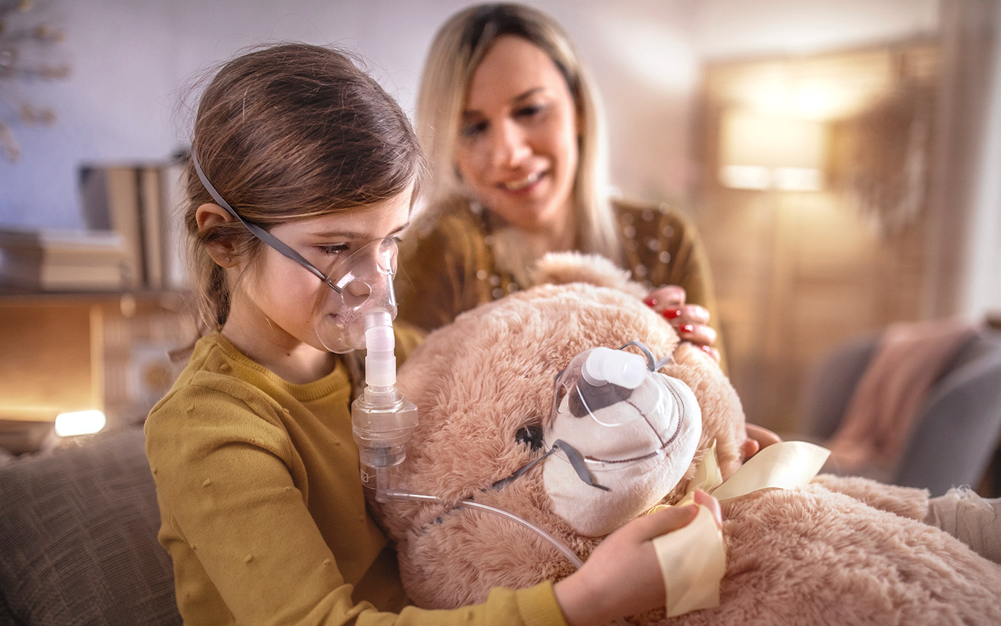 Foto: Kind mit Sauerstoff-Maske hält Teddy mit Sauerstoff-Maske im Arm. Daneben sitzt die Mutter. 