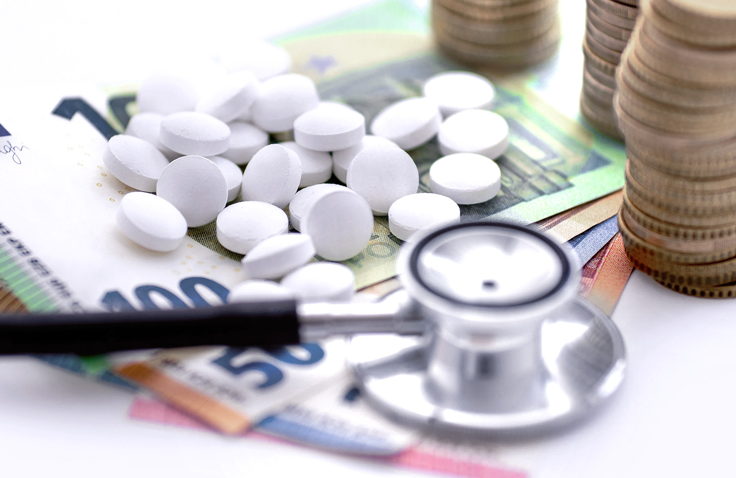 Foto: Medikamente und ein Stethoskop liegen auf Geldscheinen neben gestapelten Münzen.