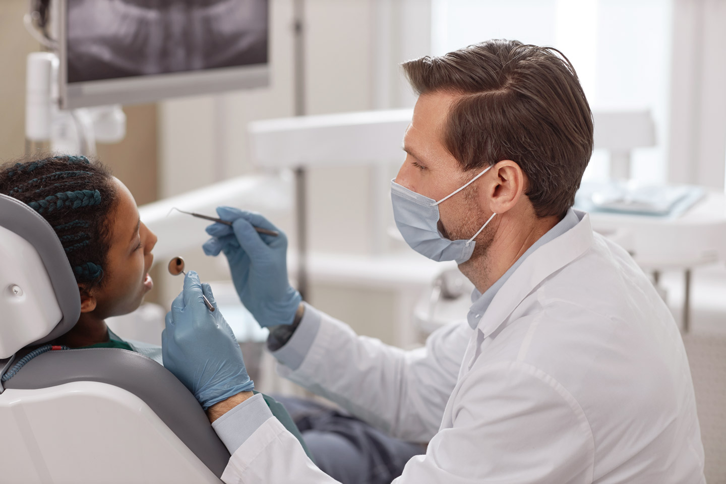 Foto: Eine Patientin sitzt mit geöffneten Mund im Behandlungsstuhl: Der daneben sitzende Zahnarzt hält einen Mundspiegel und blickt in den Mund. 