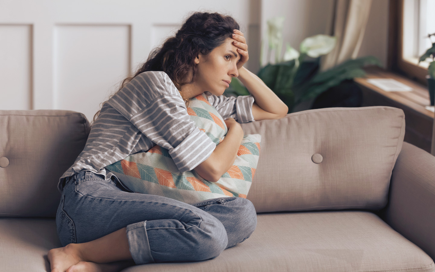 Foto: Eine Frau sitzt auf dem Sofa und schaut bedrückt und sorgenvoll vor sich hin.