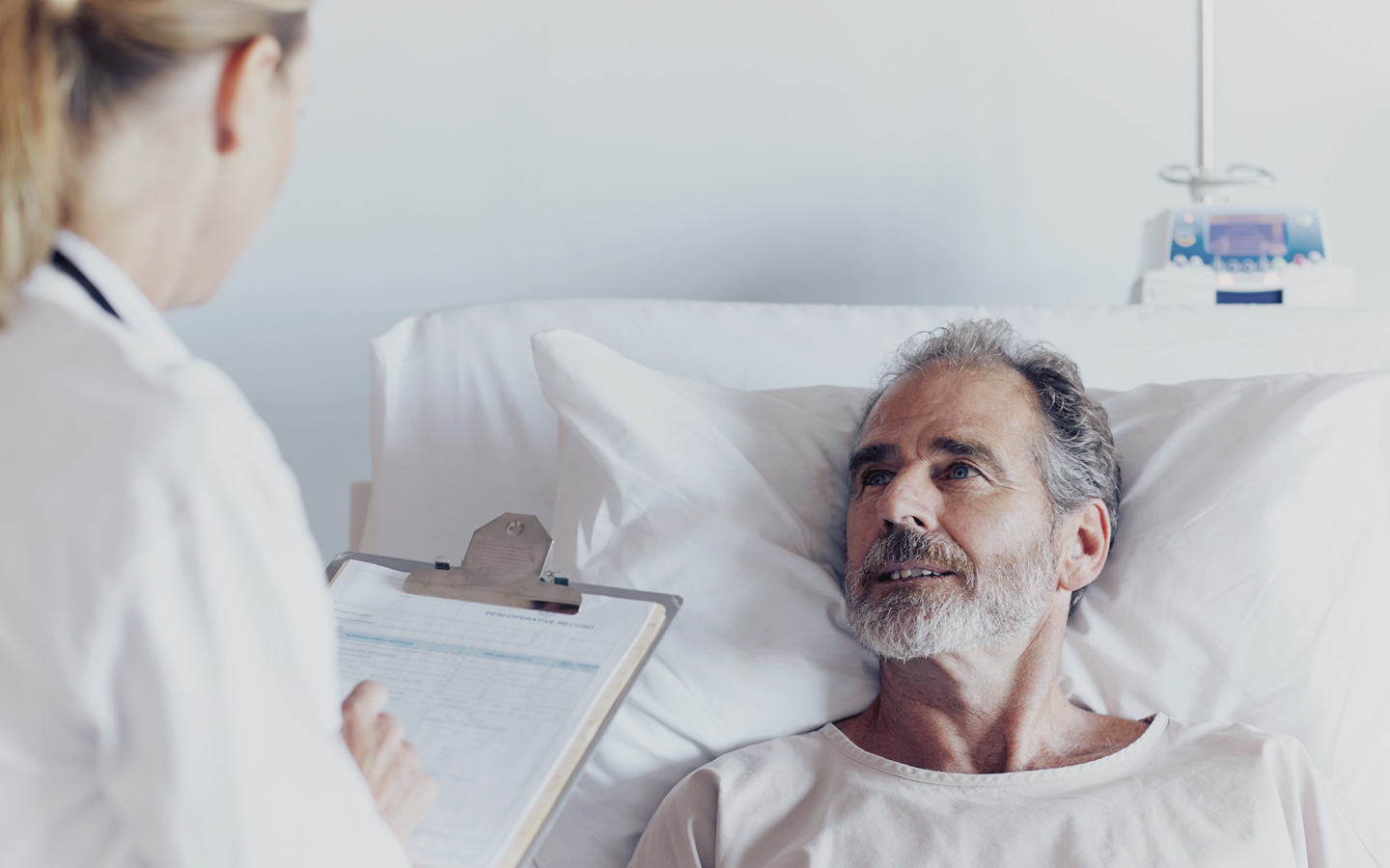 Foto: Patient liegt im Krankenhausbett und Ärztin steht daneben mit Behandlungsplan