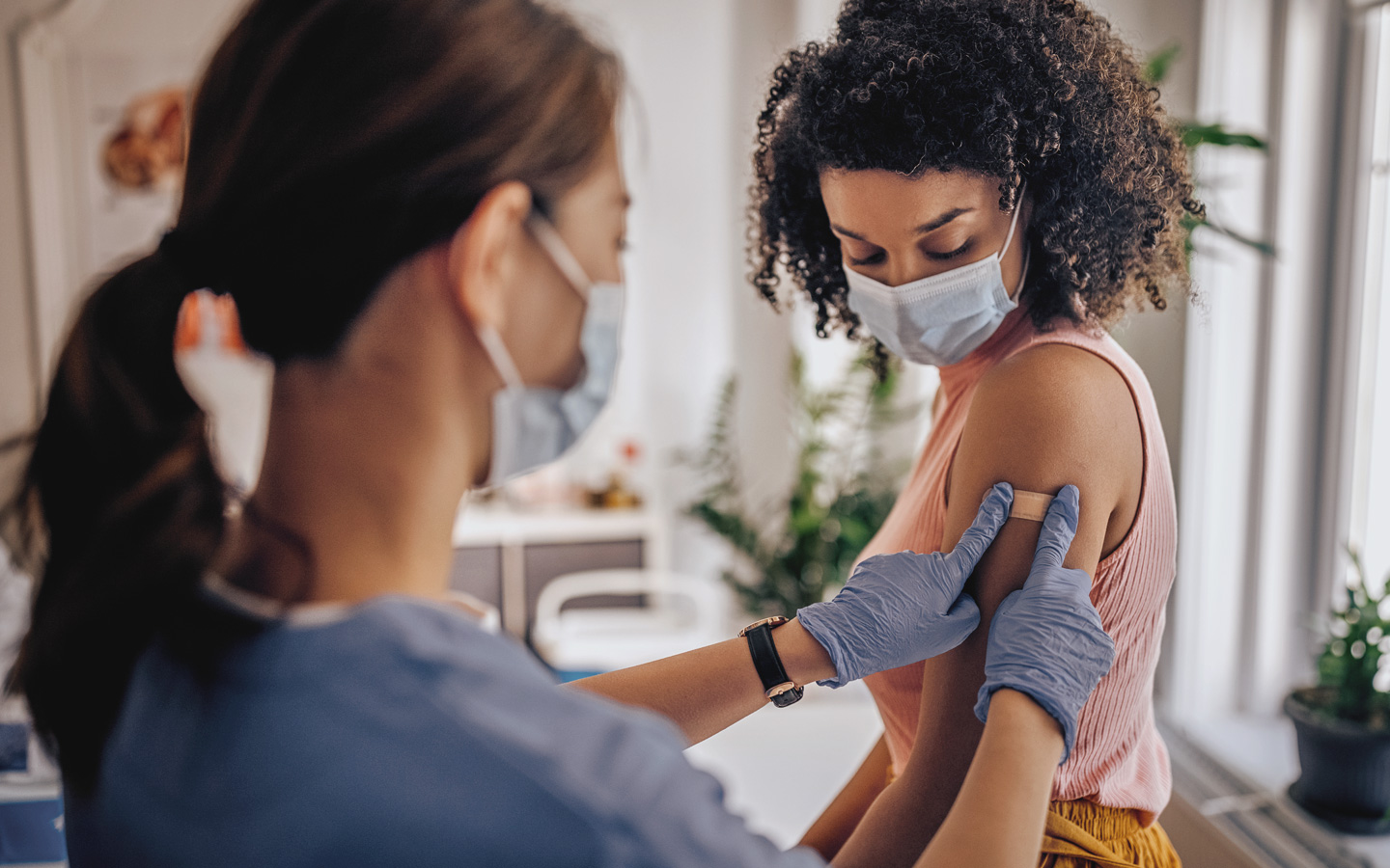 Foto: Krankenschwester mit Mundschutz und Handschuhen klebt einer Frau mit Mundschutz ein Pflaster auf den Oberarm