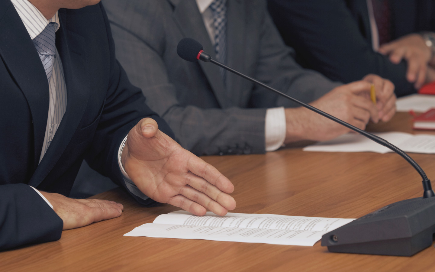 Foto: Nachaufnahme von Händen am Tisch mit Mikrofonen und Unterlagen. Eine Person im Anzug hält eine Rede und hat einen Zettel vor sich liegen mit Notizen.