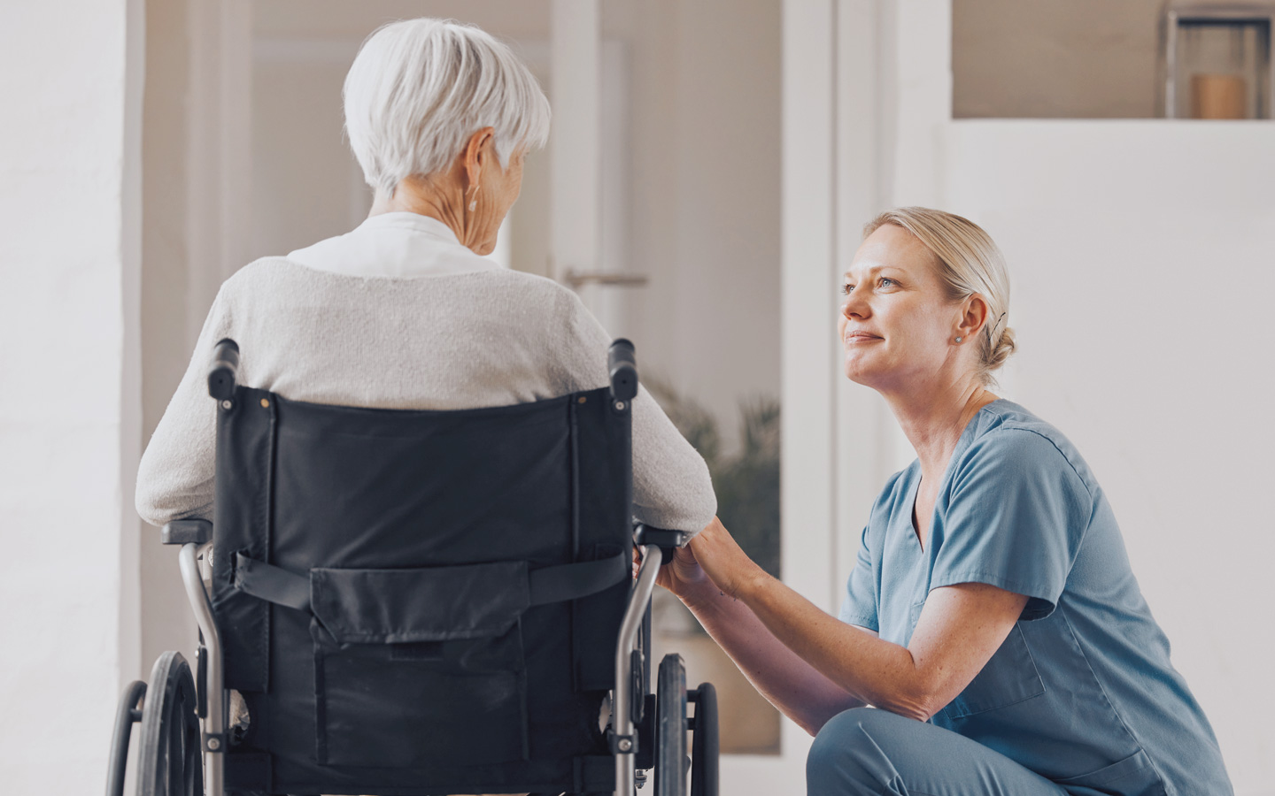Foto: Ältere Frau sitzt im Rollstuhl, Pflegerin kniet daneben und guckt sie an
